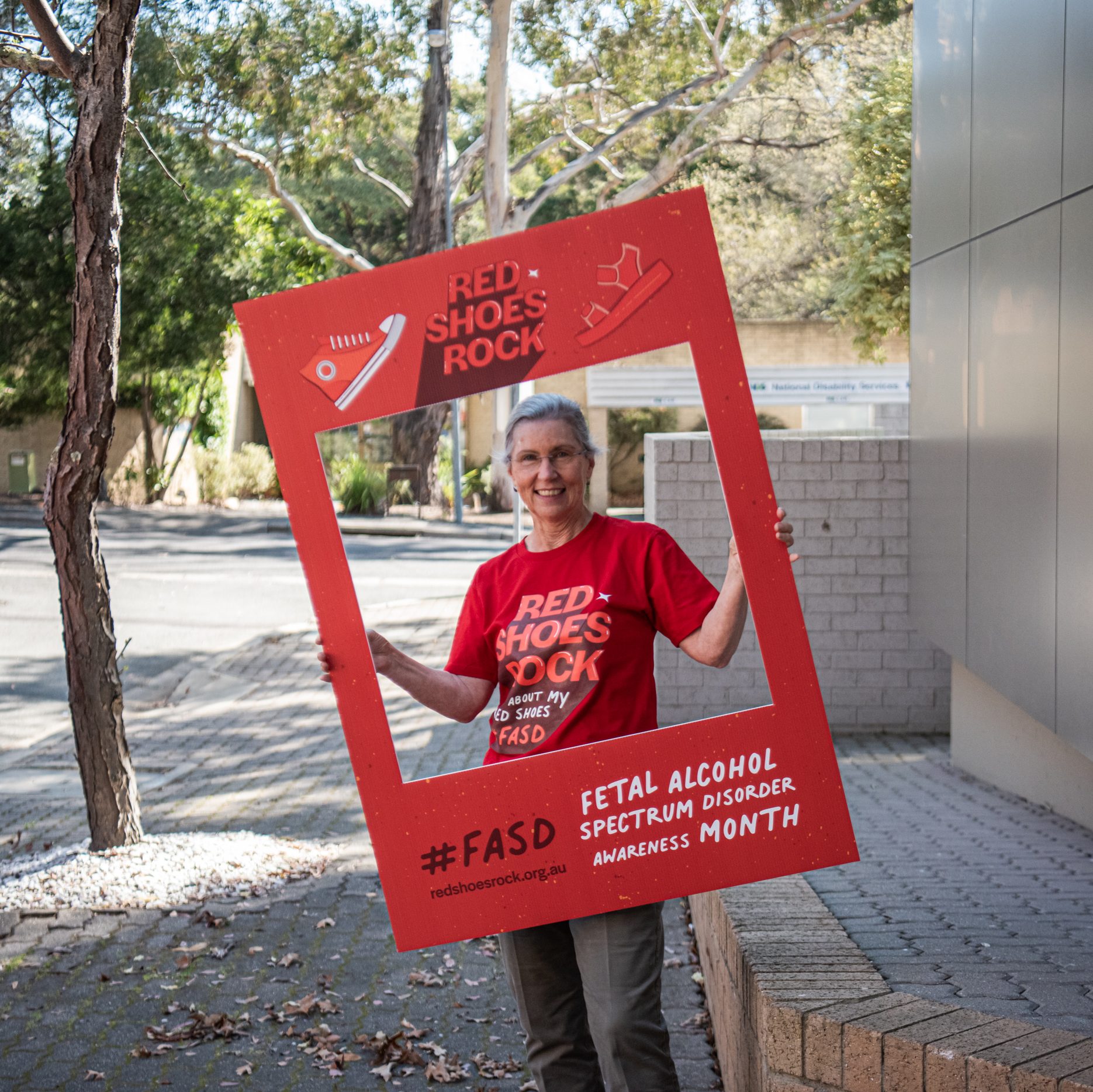 A woman standing in a red shirt, holding a banner that says 'Red Shoes Rock'.